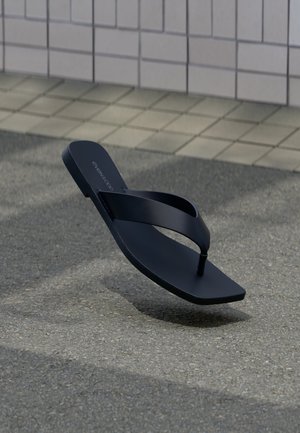 Schwarze Flip-Flop-Sandale mit Zehensteg, die über Asphalt in der Nähe eines gefliesten Gehwegs und einer Wand bei sanftem Tageslicht schwebt.