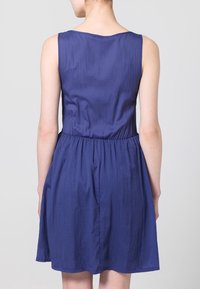 Hihaton, polven pituinen mekko, joka on valmistettu teksturoidusta, kevyestä kankaasta. Asusteena istuva vyötärö ja tummansininen väri.