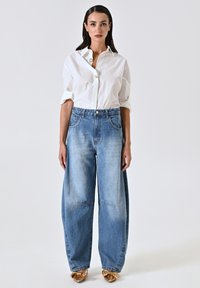 Jean large bleu avec un design délavé, associé à une chemise blanche à boutons. À noter les grandes poches et la coupe ample. Des chaussures dorées complètent le look.