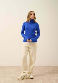 Blauer gerippter Rollkragenpullover, eng anliegendes Design, kombiniert mit crèmefarbenen weit geschnittenen Hosen und neutralen Sneakers. Einfarbiger Hintergrund.