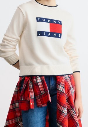 Personne portant un pull crème Tommy Jeans avec un bord bleu marine, un jean bleu et une chemise à carreaux rouge nouée autour de la taille.