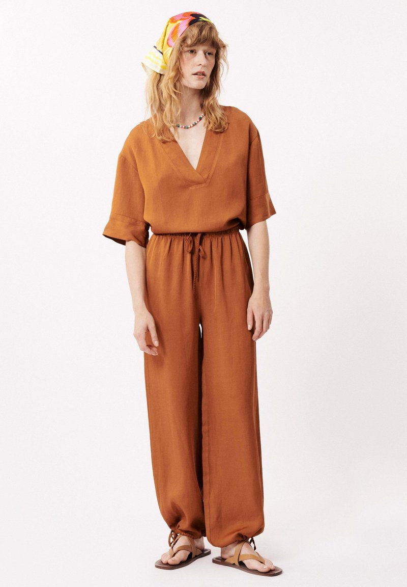 Rustkleurige jumpsuit met korte mouwen, V-hals, trekkoord in de taille en wijde benen. Gecombineerd met bruine sandalen en een meerkleurige hoofddoek.