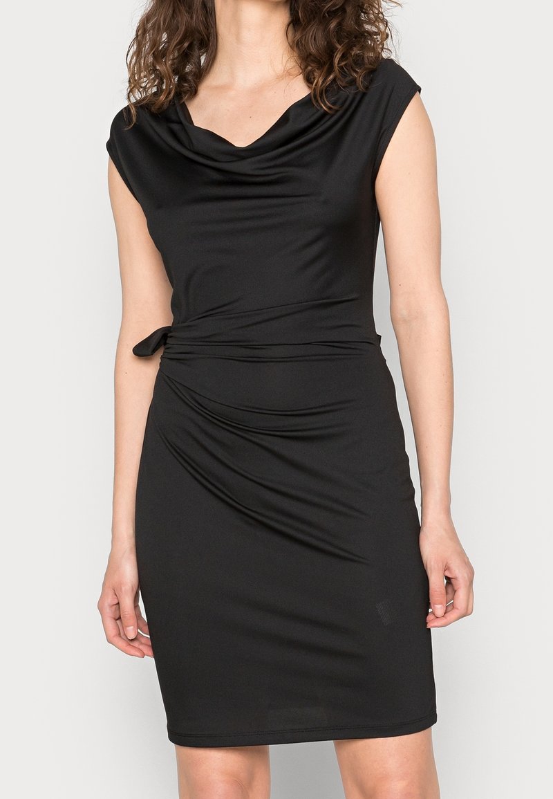 Femme portant une robe noire sans manches ajustée avec un décolleté drapé et un détail noué sur le côté, debout devant un fond clair uni.