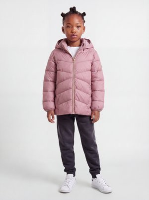 Jeune fille debout portant une doudoune mauve, un pantalon noir, des baskets blanches et une chemise blanche sur fond blanc.