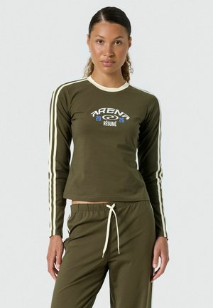 Žena oblečená v olivovozelenej dlhorukávovej blúzke a zodpovedajúcich nohaviciach s bielymi pruhmi a nápisom "Arena Résumé 2026" na tričku.