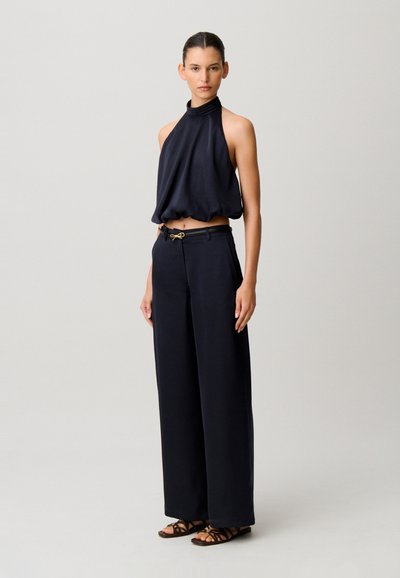 Donna che indossa un top nero senza maniche con collo all'americana e pantaloni neri a gamba larga e vita alta, con sandali, in piedi davanti a uno sfondo chiaro uniforme.