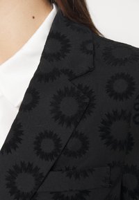 Veste noire avec un motif floral texturé, col à revers et détail de poche. En dessous, une chemise blanche à col est partiellement visible.