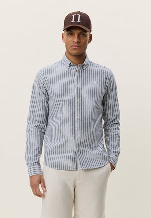 Man som bär en brun keps och en blåvit vertikalt randig skjorta med knappar och beige byxor, stående mot en enfärgad bakgrund.