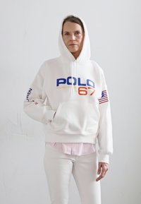 Balta kapuce ar "POLO 67" zilu un orandžu drukājumu, kabata kangarū, un Amerikas karoga uzpleca. Valkāta pār rozā svītrainu krekls.