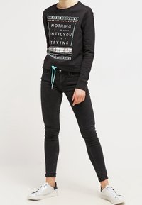 Sudadera negra con impresión de texto y detalles estampados, combinada con jeans negros ajustados y zapatillas blancas con acentos negros.