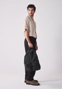 Svart nylonryggsäck med två dragkedjefickor, hålls i en hand. Modellen bär en beige kortärmad skjorta och svarta byxor med bruna kängor.