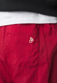 Pantaloni corti rossi realizzati in un tessuto leggero, con una tasca posteriore con logo ricamato beige. La texture è leggermente sgualcita.