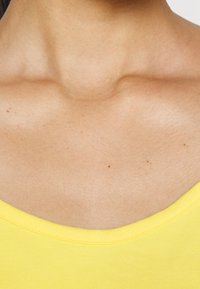 Gul linne med rundad halsringning, med en slät textur och minimala sömdetaljer, vilar mot bar hud.