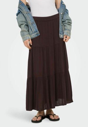 Femme portant un débardeur blanc, une veste en jean tombant des épaules, une longue jupe à volants foncée et des sandales noires à brides, debout sur un fond uni.