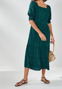Tiefgrünes Midikleid mit schwarzen Punkten, viereckigem Ausschnitt, Puffärmeln und mehrlagigem Rüschensaum, kombiniert mit braunen Riemchensandalen und einer geflochtenen Tasche.