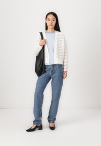 Cárdigan blanco de punto con tejido abierto sobre una camisa con rayas azules, combinado con jeans azul claros y zapatos de tacón negros, llevando un bolso negro.
