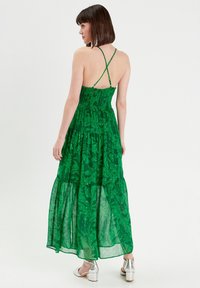 Groene maxi-jurk met een textuur in het lijfje, kruisbanden op de rug en gelaagd, doorschijnend stof met een bladerenpatroon. Gecombineerd met zilveren chunky hakken.