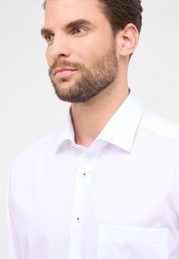 Camicia bianca realizzata in tessuto liscio, con colletto classico, una tasca sul petto e bottoni scuri sulla pattina.