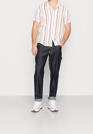 Kortemouwshirt in het wit met rode verticale strepen, gecombineerd met donkerblauwe jeans. Witte sneakers met lichtbruin accent en opgerolde mouwen.