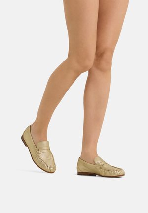 Woman’s legs wearing metallic gold slip-on loafers with low wooden heels against a plain light background.