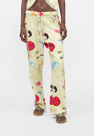 Vrouw draagt losse lichtgele broek met kleurrijke maritieme en bloemmotieven, staat op blote voeten met iets gespreide benen.