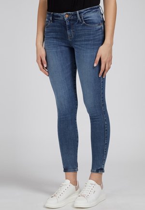 Kobieta ubrana w niebieskie damskie dżinsy z średnim stanem i białe trampki, stojąca na tle jednolitego jasnoszarego tła.