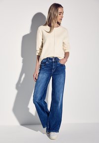 Weite blaue Denim-Jeans kombiniert mit einem hellbeigen Rundhals-Pullover. Die Jeans verfügen über ein klassisches Fünf-Taschen-Design und eine subtile Ausbleichung.