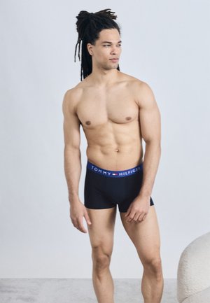 Homme musclé avec de longues dreadlocks portant un boxer noir Tommy Hilfiger, debout devant un fond clair uni.