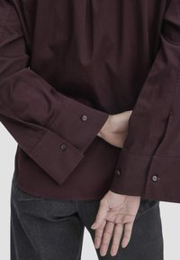 Camicia button-up bordeaux con maniche lunghe, caratterizzata da due polsini con bottone, realizzata in tessuto liscio. Indossata con jeans scuri.