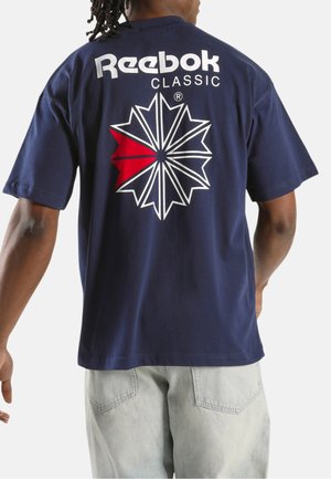Mann trägt ein dunkelblaues Reebok Classic T-Shirt mit einem weißen und roten Sternexplosions-Logo auf dem Rücken und hellen Jeans.
