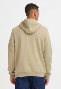 Sudadera con capucha beige de corte relajado, con puños y dobladillo de canalé, que presenta una textura suave y una etiqueta de logo discreta en el lateral.