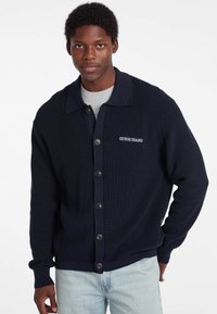 Námornícky pletený cardigan s golierom, zapínaním na gombíky a vyšitým logom "GUESS JEANS". Mäkko struktúrovaný, s dlhými rukávmi, vo voľnom strihu.