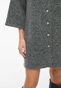 Pilka vilnos mišinys paltas su tekstūruotu paviršiumi, per dideli rankovės ir penki metaliniai mygtukai priekyje. Pasižymi tiesiu apvadu.