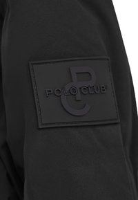 Svart jacka med en texturerad, svart gummipatch med präglingen "POLO CLUB" och monograminitialer. Slät tyg, strukturerad design.