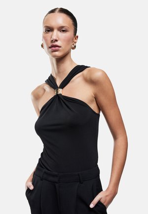 Femme portant un haut noir sans manches avec un fermoir doré et un pantalon noir assorti, mains dans les poches, expression neutre, fond clair.