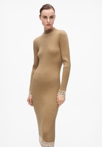 Abito a maglia a maniche lunghe a coste in beige con colletto alto. Presenta il logo in contrasto "LAGERFELD" su polsini e orlo. Silhouette aderente.