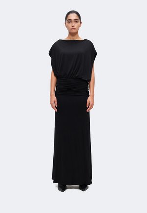 Femme aux cheveux foncés dans une longue robe noire à taille froncée, debout droite devant un fond clair uni.