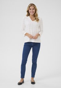 Haut blanc à manches longues avec motif en œillets, décolleté en V, coupe relaxée, associé à un jean skinny bleu et des ballerines noires, se tenant sur un fond uni.