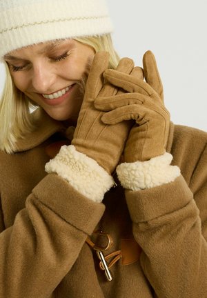Gants en suède marron avec des poignets en shearling crème, texture lisse et coutures détaillées, portés par une personne dans un manteau marron et un chapeau blanc.