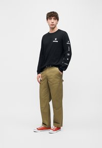 Νεαρός άνδρας όρθιος, φοράει μαύρο μακρυμάνικο πουκάμισο Converse, χακί παντελόνι cargo και κόκκινα sneakers χαμηλού κόψιμο, μπροστά σε απλό λευκό φόντο.