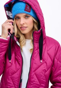 Fuchsiová prošívaná bunda s kapucí, s černými stahovacími šňůrkami a zipovým zapínáním. Nosič má na sobě modrou pletenou čepici s vzorovou podšívkou.