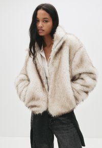 Kurtka z sztucznego futra w jasnym beżu o puszystej fakturze, w oversize'owym kroju i z zapięciem na zamek. Noszona na białej koszuli i czarnych dżinsach.