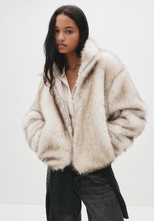 Veste en fausse fourrure beige clair avec une texture pelucheuse, coupe oversize et fermeture éclair. Portée par-dessus une chemise blanche et un jean noir.