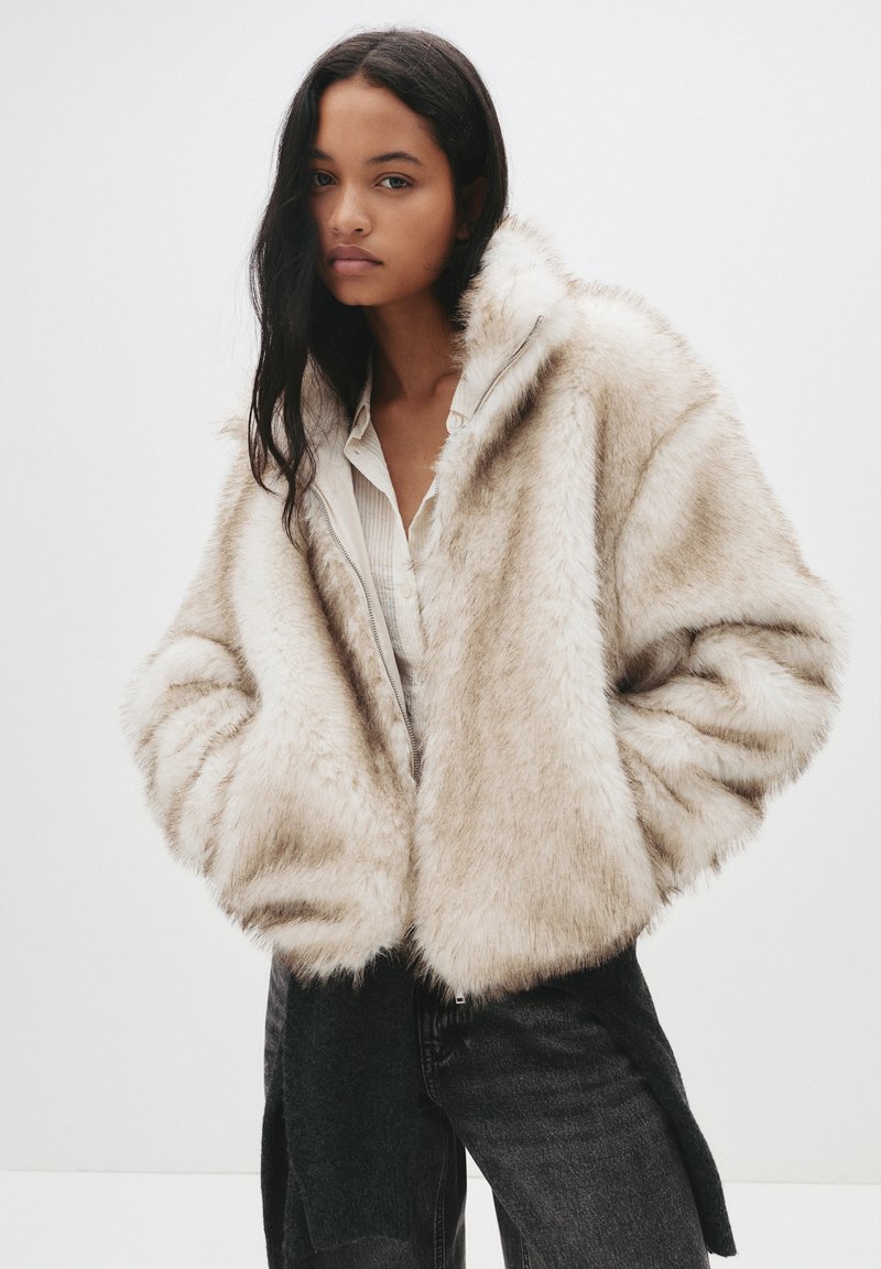 Kurtka z sztucznego futra w jasnym beżu o puszystej fakturze, w oversize'owym kroju i z zapięciem na zamek. Noszona na białej koszuli i czarnych dżinsach.
