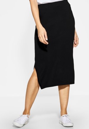 Kvinde iført en sort knælange nederdel med en sideslids og hvide sneakers, stående foran en ensfarvet hvid baggrund.