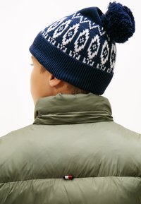 Veste matelassée verte avec un col haut et une petite étiquette de marque, associée à un bonnet tricoté bleu à motif, surmonté d'un pompon.