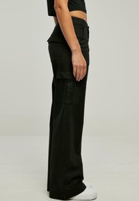 Vue de profil d'une personne portant un pantalon cargo noir à jambes larges avec des poches à rabat et des baskets blanches sur un fond uni.