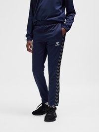 Persoon draagt marineblauwe Hummel trainingsbroek met chevronpatroon aan de zijkanten en zwarte sneakers, staand met één hand in de zak.