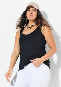 Top negro sin mangas con cuello en V, hecho de tela suave, combinado con pantalones blancos; el modelo lleva una gorra de color claro y joyas llamativas.