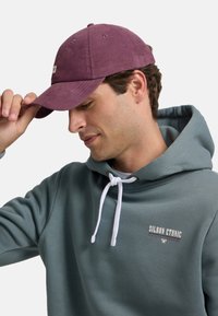 Gorra de pana púrpura con parche bordado, combinada con una sudadera con capucha verde azulado que cuenta con cordones blancos y detalle de logo.